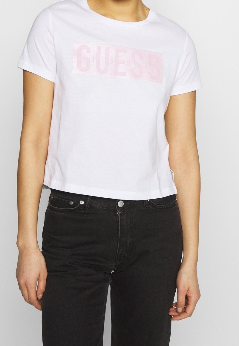 Hvid cropped t-shirt lavet af bomuld med korte ærmer, prydet med et blankt lyserødt "GUESS" logo på forsiden. Kombination med sorte jeans.