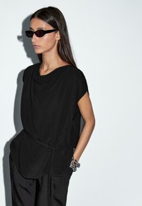 Top nero con scollatura drappeggiata, maniche corte e lacci laterali. La modella indossa occhiali da sole neri eleganti e un braccialetto decorativo bianco e nero.