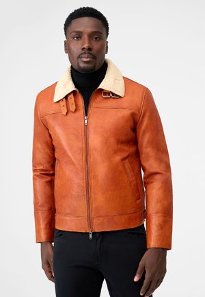 Mann trägt eine hellbraune Lederjacke mit cremefarbenem Lammfellkragen über einem schwarzen Rollkragenpullover und schwarzen Hosen vor einem schlichten Hintergrund.