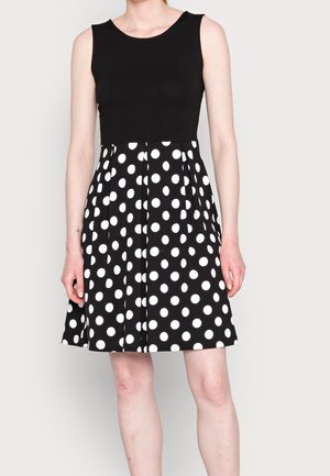 Vrouw draagt een mouwloze zwarte jurk met een witte rok met stippen, staand tegen een eenvoudige lichtgrijze achtergrond.