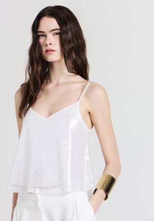 Top corto bianco con scollo a V, tessuto trasparente e dettagli in paillettes. Indossato con un bracciale a manica texturizzato dorato.