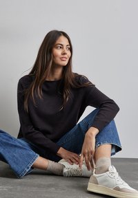 Maglione nero a maglia con scollo rotondo, abbinato a jeans a gamba larga di un blu chiaro e sneaker bianche con dettagli beige, seduto su un pavimento grigio.