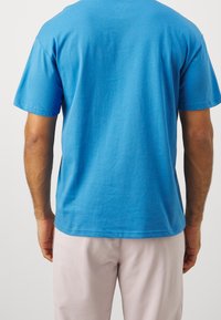 Blue cotton t-shirt with short sleeves and a crew neck, viewed from the back. Smooth texture, casual fit, and stitched hem details.