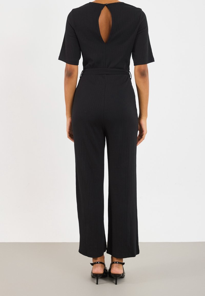 Combinaison noire côtelée avec manches courtes, décolleté dos ouvert, taille ajustée et pantalons à jambes larges. La texture semble lisse.