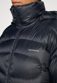 Črna puff jakna z visokim ovratnikom, prešita zasnova, z zadrgo spredaj in majhnim logotipom "SALOMON" na prsih. Gladek, lahek material.