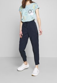 Lichtblauw T-shirt met cirkelpatronen, gecombineerd met marineblauwe joggers met elastische taille en met rits afgesloten zakken. Witte sneakers.