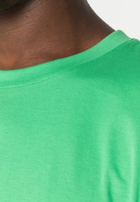 T-shirt verde in cotone con scollo rotondo, caratterizzato da un tessuto liscio e leggermente texturizzato e cuciture visibili lungo il colletto.
