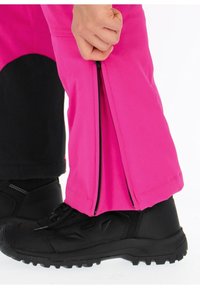 Roze buitensp trousers met een zijritsfunctie, gemaakt van flexibel materiaal, gecombineerd met een zwarte boot op een gestructureerd oppervlak.