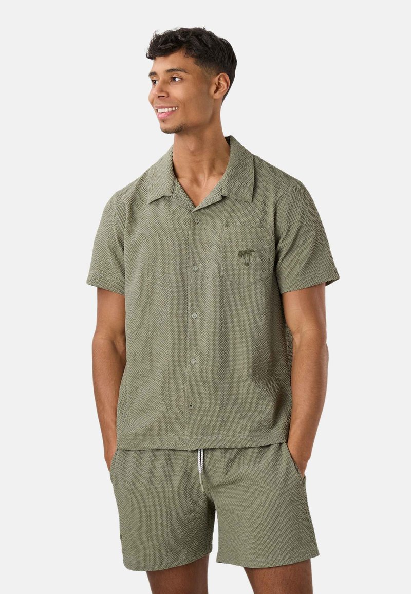 Jeune homme souriant, portant une chemise à manches courtes texturée vert olive avec boutons et un short assorti, les mains dans les poches.