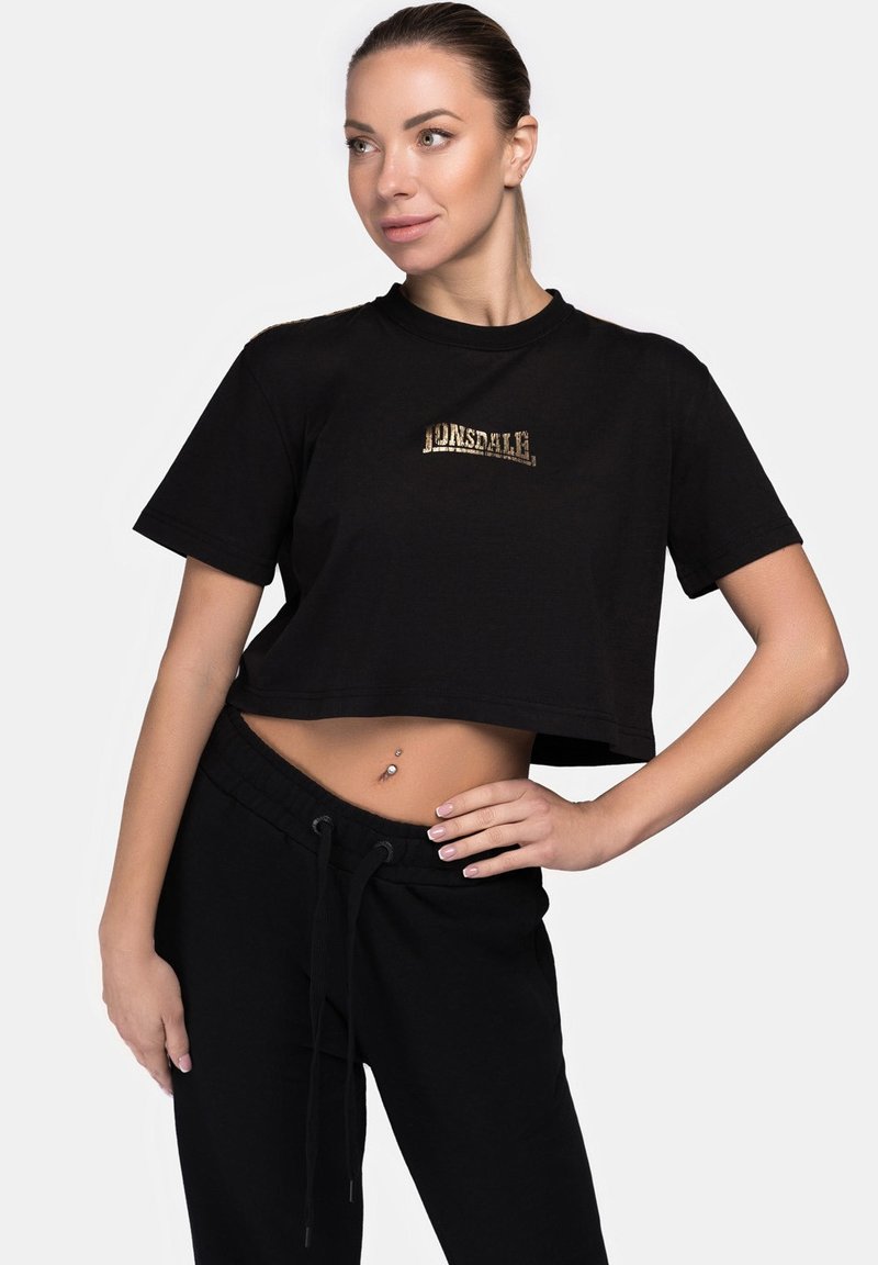 Fekete, pamutból készült, rövid ujjú, arany Lonsdale logós cropped póló, kerek nyakkivágással. A modellen fekete nadrág van.