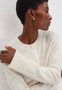 Frau mit kurzen, lockigen Haaren, die silberne Ohrringe und eine crema-farbene Strickjacke mit großen Knöpfen trägt, schaut zur Seite mit verschränkten Armen.