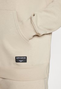 Sweatshirt bege com uma textura macia, apresentando um bolso frontal e um emblema preto com texto branco, localizado na parte inferior esquerda.