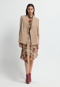 Vrouw in een beige dubbelzijdige blazer over een bruin gemarmerde midi-jurk, gecombineerd met bordeauxrode enkellaarsjes, staand tegen een witte achtergrond.