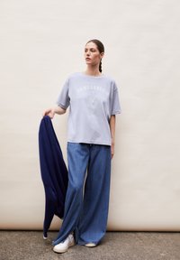 Lichtblauw oversized t-shirt met hartprint, gecombineerd met een wijde denim broek. Houdt een marineblauwe jas vast, gestyled met witte sneakers.