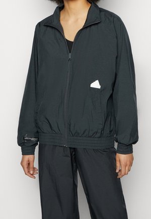 Persona vistiendo una chaqueta negra con cremallera, con puños y dobladillo elásticos, combinada con pantalones negros a juego, de pie frente a un fondo liso.