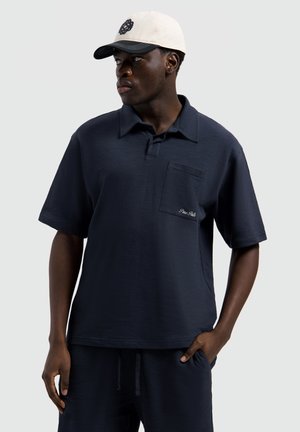 Man met een marineblauw shirt met korte mouwen en borstzak, en een witte pet, kijkend naar de zijkant met één hand in de zak.