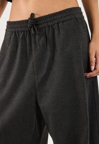 Mørkegrå sweatpants med elastisk talje og snøre, båret af en person, der viser maven og har højre hånd hvilende nær lommen.