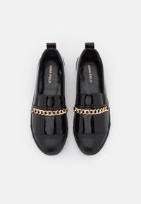 Svarta lackskinnsskor i slip-on-stil med en guldkedja som accent över framsidan. Har en slät yta och en rundad tådesign.