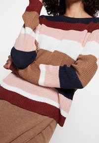 Stickad tröja med horisontella ränder i rosa, vitt, brunt och marinblått. Rutig form med ribbade ärmslut och nederkant, som visar ett texturerat stickat mönster.