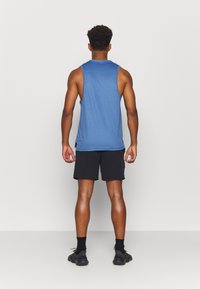 Man standing facing away wearing a blue sleeveless shirt, black shorts, black socks, and black athletic shoes against a plain background.