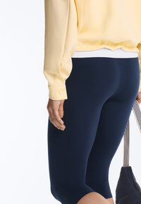 Gros plan d'une personne portant un t-shirt jaune et des leggings bleu marine, tenant un sac noir avec des poignées rayées.