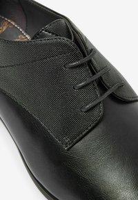 Zapato de cuero negro con parte superior de lienzo texturizado, acabado brillante suave y detalles de costura sutiles, con tres ojetes para los cordones.