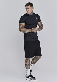 Melns pieguļošs īsām piedurknēm krekls, melni šorti un melnas sporta apavi ar baltiem akcentiem. Izteiksmīgas tetovējumi uz rokām un kājām.