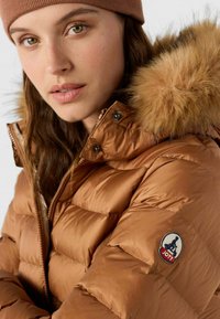 Bruine donzen jas met een door bont gevoerde capuchon, voorzien van een doorgestikt ontwerp en een logopatch op de mouw. Gladde textuur met een glanzende afwerking.