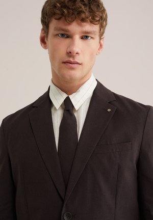 Mladi moški s kodrastimi lasmi, oblečen v temno obleko, črno kravato in belo srajco s ovratnikom, na enostavni ozadju.