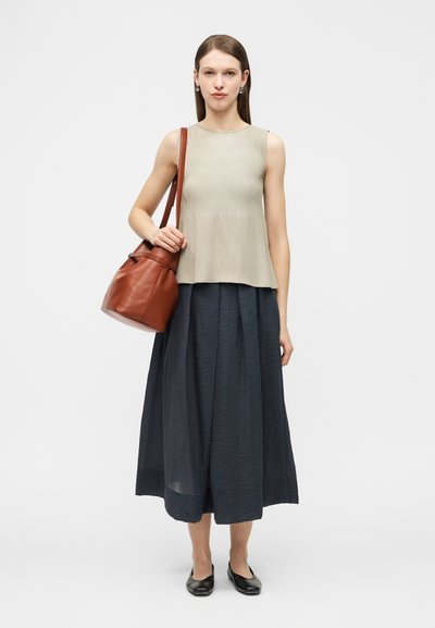 Femme debout portant un haut beige sans manches, une jupe midi bleu foncé, des ballerines noires, et un grand sac à bandoulière en cuir marron.