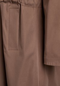 Detal ubrania z brązowego materiału, z płaską kieszenią, gładką teksturą i prostym wzornictwem, z widocznym przeszyciem wzdłuż szwów.