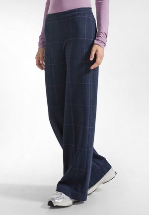 Persona que lleva pantalones de pierna ancha azul marino con un patrón de cuadros morados y zapatillas deportivas plateadas, combinados con una camiseta de manga larga color púrpura claro.