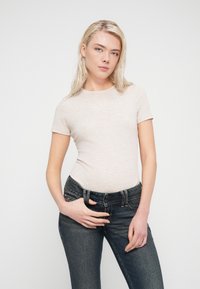 Φωτεινό μπεζ εφαρμοστό μπλουζάκι με κοντά μανίκια, κατασκευασμένο από απαλό υλικό, σε συνδυασμό με σκουρόχρωμα τζιν παντελόνια με κλείσιμο με κουμπί και τσέπες.