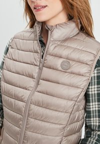 Hellbeige Puffer-Weste mit gestepptem Design, hohem Kragen und Reißverschluss vorne. Akzentuiert mit einem runden Logopatch auf der Brust.