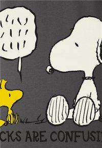 Sort t-shirt med Snoopy og Woodstock illustrationer. Teksten lyder: "STICKER ER FORVIRRENDE." Enkel, tegneserieagtig stil med klare farver.