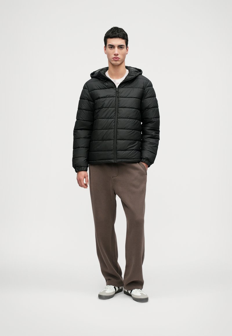 Veste matelassée noire avec capuche, fermeture éclair à l'avant et design matelassé. Associée à un pantalon de survêtement marron et des baskets blanches avec des rayures noires.