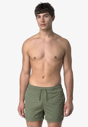 Giovane uomo in piedi, senza maglietta, indossa pantaloncini da bagno verde oliva con coulisse su uno sfondo chiaro uniforme.