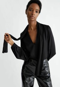 Blusa nera con scollatura a V e maniche fluide, abbinata a pantaloni neri lucidi. Una sciarpa plissettata si appoggia sulla scollatura, aggiungendo texture.