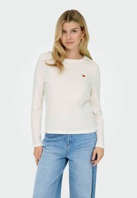 Hvid langærmet t-shirt lavet af blødt stof, med et lille rødt hjerte broderet på brystet, parret med lyseblå brede jeans.