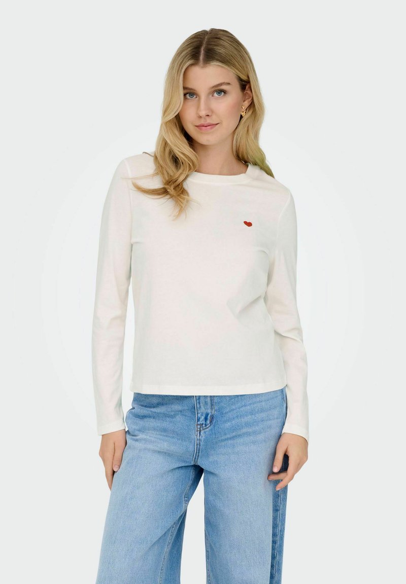 Hvid langærmet t-shirt lavet af blødt stof, med et lille rødt hjerte broderet på brystet, parret med lyseblå brede jeans.
