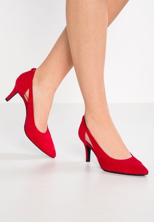 Jambes portant des chaussures slingback en daim rouge à talons bas, debout sur un sol blanc sur fond uni.