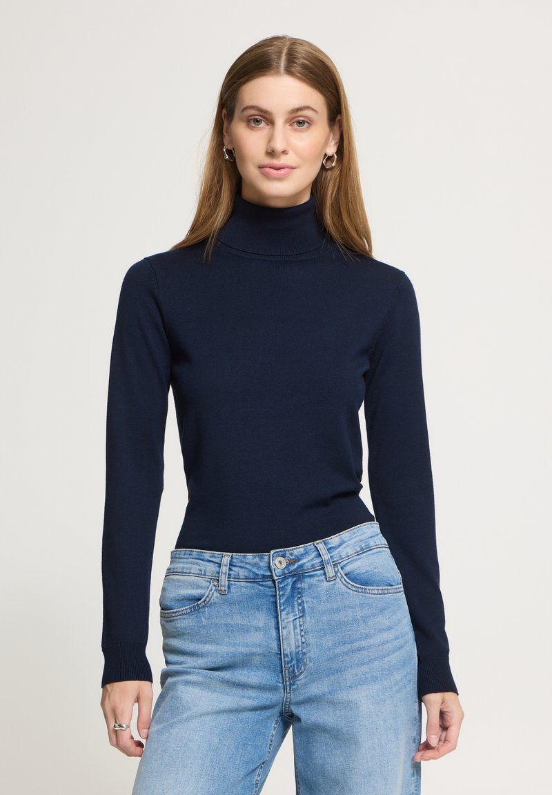 Femme debout, portant un pull à col roulé bleu foncé et un jean bleu clair, de face devant un fond uni clair.