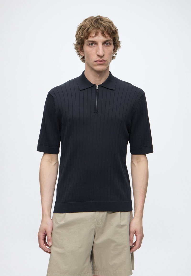 Jeune homme aux cheveux châtain clair bouclés porte un polo noir à manches courtes côtelé et un short beige sur un fond clair uni.