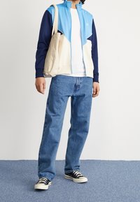 Sportjacka i ljusblått och marinblått, med huva och dragkedja, kombinerad med en vit t-shirt och blå jeans, samt svarta sneakers. Beige tygkasse.