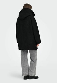 Črn oversize volnen plašč s kapuco, z teksturirano površino in spušenimi rameni, v kombinaciji s sivimi širokimi hlačami in črnimi čevlji.