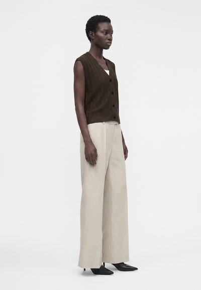 Femme debout portant un gilet sans manches marron foncé boutonné, un pantalon large beige et des talons noirs à bout pointu sur un fond uni.