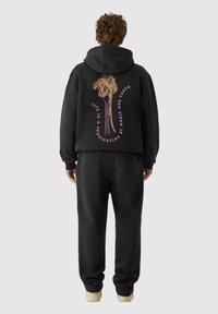 Set di felpa con cappuccio e pantaloni neri. Sul retro è presente una grafica di spaghetti in giallo e rosa con la scritta: "La vita è una combinazione di magia e pasta."