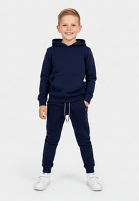 Granatowa bluza z kapturem z przednią kieszenią oraz pasujące dresy z regulowanym paskiem w talii, połączone z białymi tenisówkami. Gładki materiał, luźny krój.