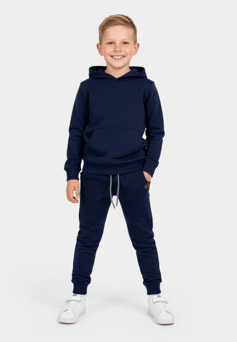 Granatowa bluza z kapturem z przednią kieszenią oraz pasujące dresy z regulowanym paskiem w talii, połączone z białymi tenisówkami. Gładki materiał, luźny krój.