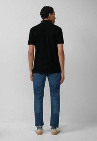 Schwarzes Polohemd mit kurzen Ärmeln, Kunststoffkragen, kombiniert mit blauen Jeans und weißen Sneakern; schlichte Rückansicht vor einem neutralen Hintergrund.